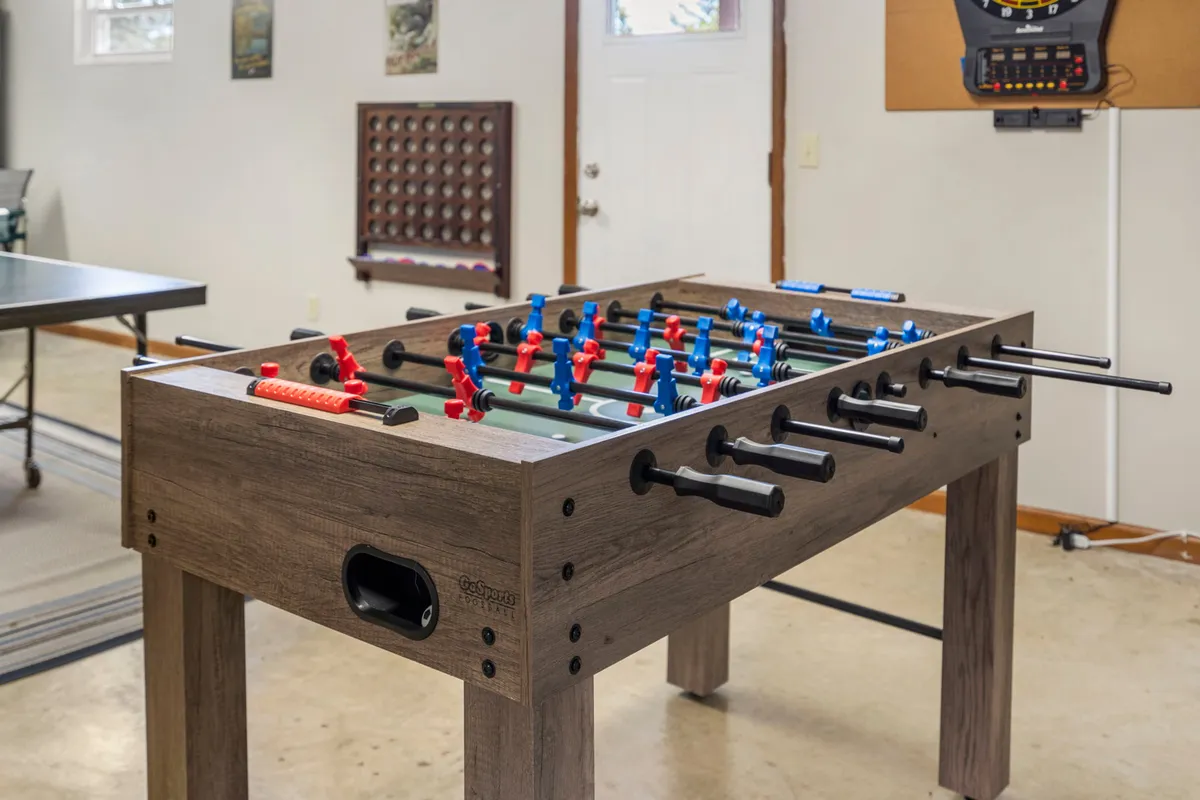 A lively interior shot of the garage game room featuring a foosball table, an electronic dart board mounted on the wall, and a ping pong table. The space is bright and energetic, clearly set up for fun and friendly competition among family members.