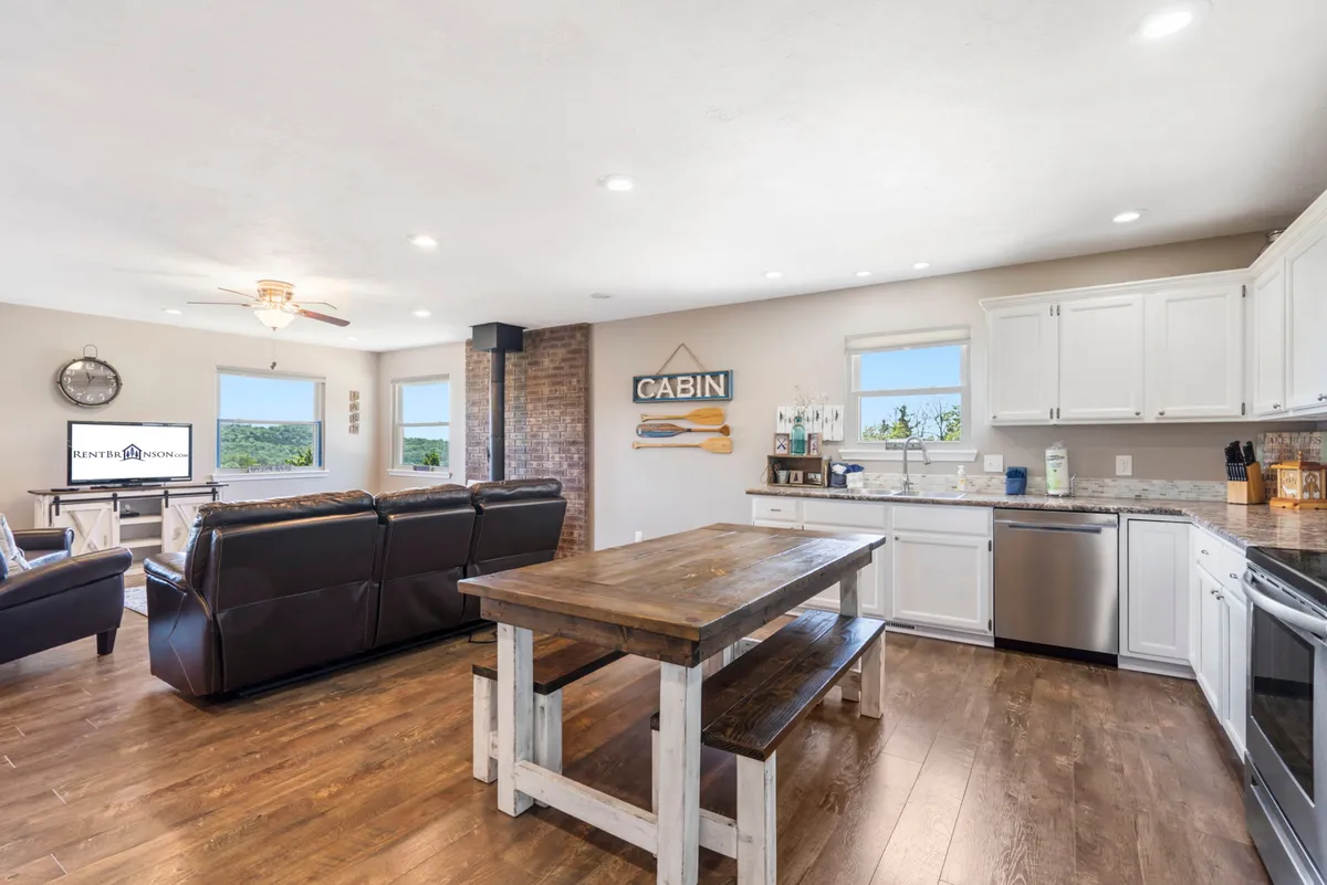 A spacious, open-concept layout featuring a farmhouse-style dining table in the foreground, a comfortable sofa, and a kitchen area visible in the background. The space is well-lit, inviting, and clearly designed for family meals, movie nights, and social gatherings.