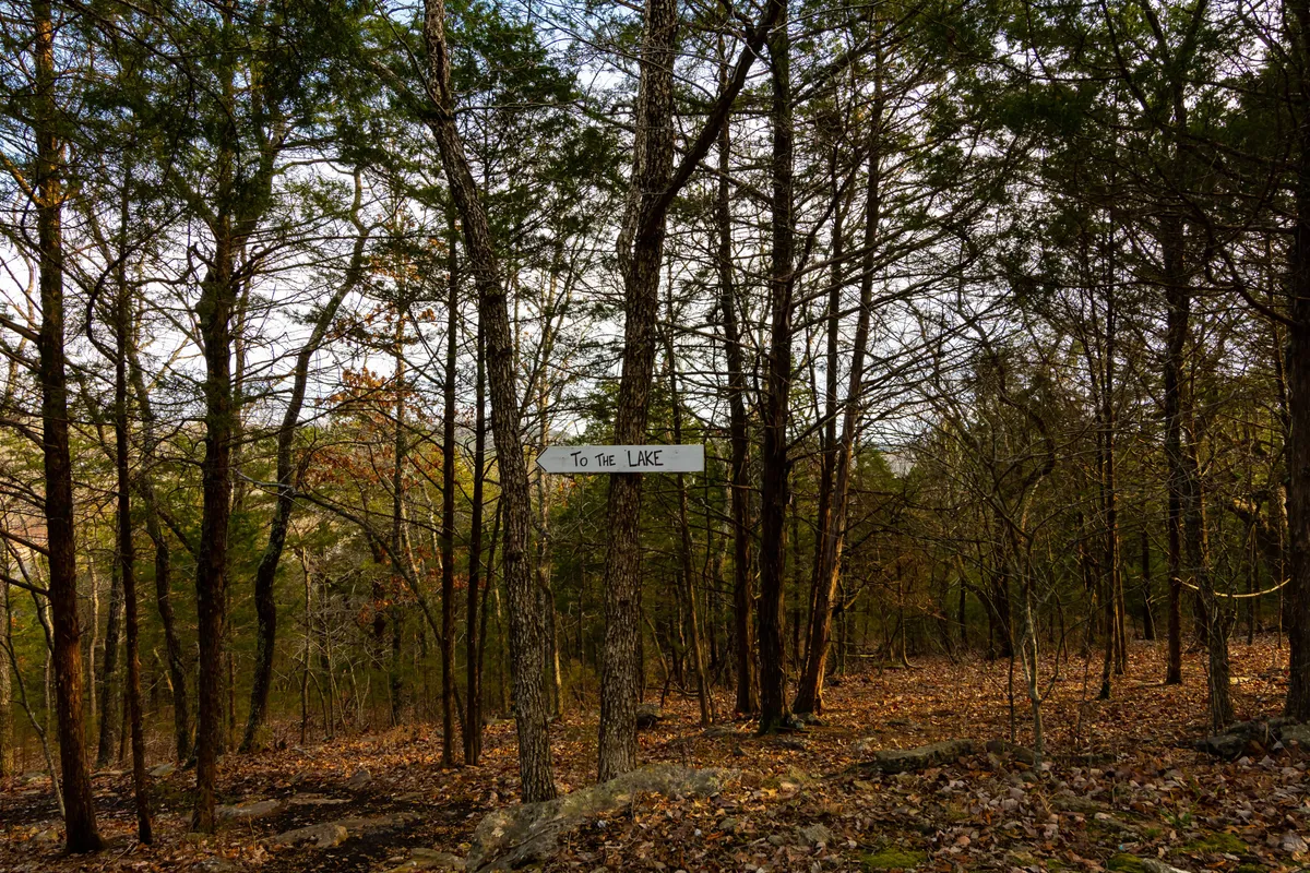 A wooded pathway marked by a sign pointing toward the lake, with trees and a leafy ground setting the scene. The trail leads guests to private lakeside access, offering moments of peaceful exploration and connection with nature.