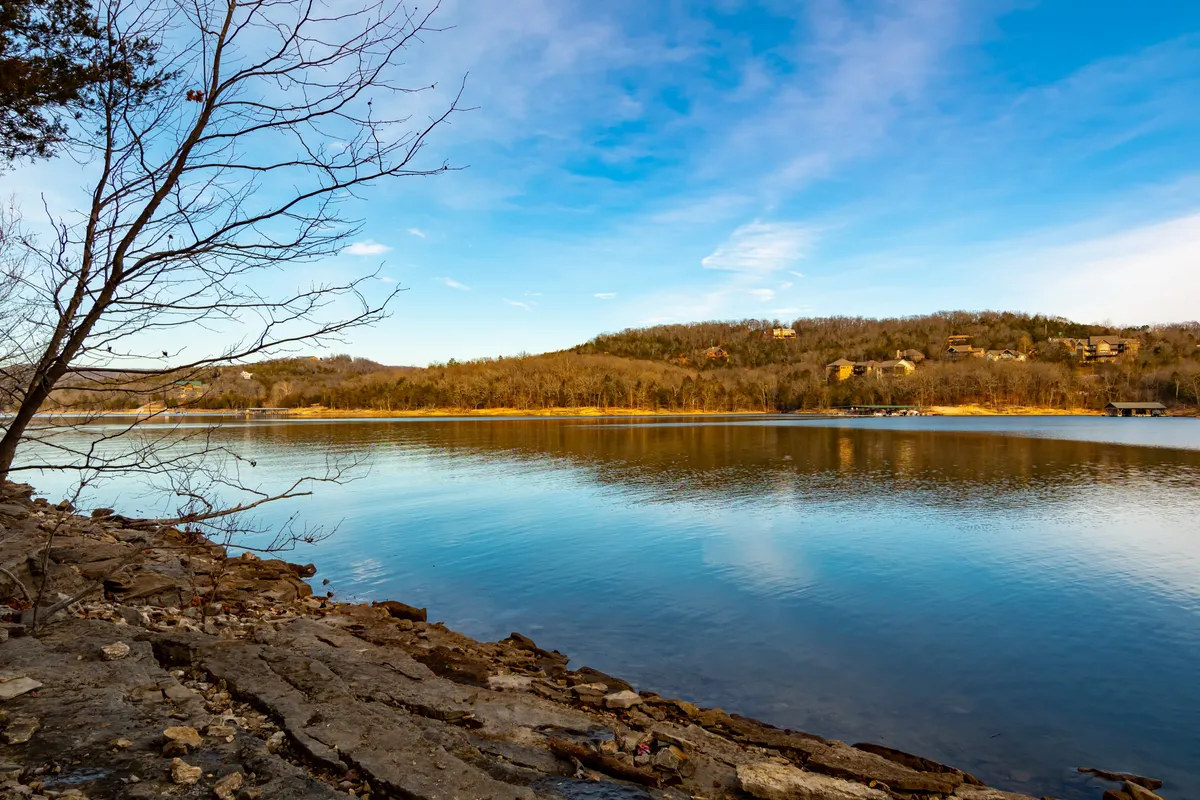 A tranquil view of the calm lake surface framed by bare tree branches, with distant hills rising in the background. The natural scenery invites quiet reflection and outdoor adventure.