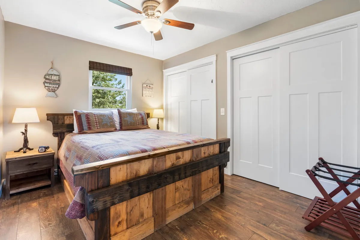 A serene bedroom with a wooden queen bed frame, soft lighting from a table lamp, and a window with a relaxing view. Wall decor and cozy bedding create a tranquil retreat ideal for restful sleep or quiet alone-time.