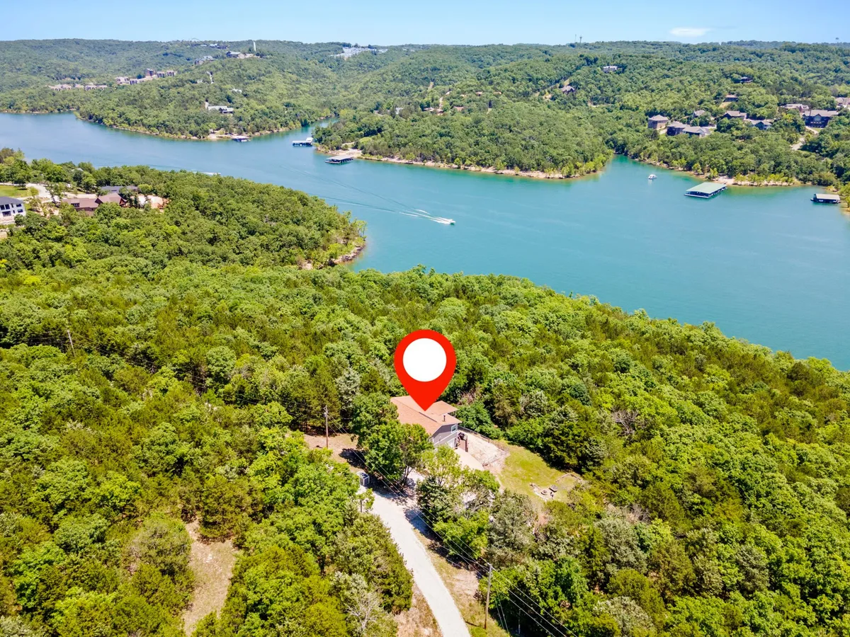 A wide view of the lake framed by lush trees, with the house and driveway visible in the foreground. The scene is tranquil, showing the proximity of the home to the water and the surrounding natural landscape, ideal for nature walks or lakeside relaxation.