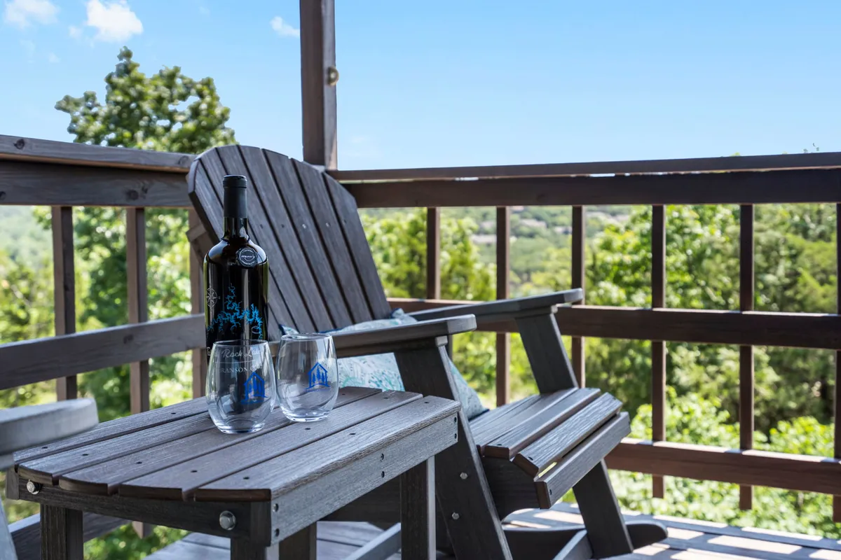 A serene porch scene with a wooden chair, a wine bottle, and glasses, all set against a backdrop of lush greenery. The setup invites guests to relax outdoors and enjoy the tranquil environment.