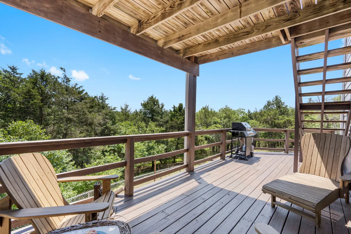 A spacious wooden deck outfitted with comfortable outdoor seating, a gas BBQ grill, and stairs leading down to the surrounding greenery. The deck offers a tranquil spot for meals, gatherings, or simply enjoying the view of nature and the lake in the distance.